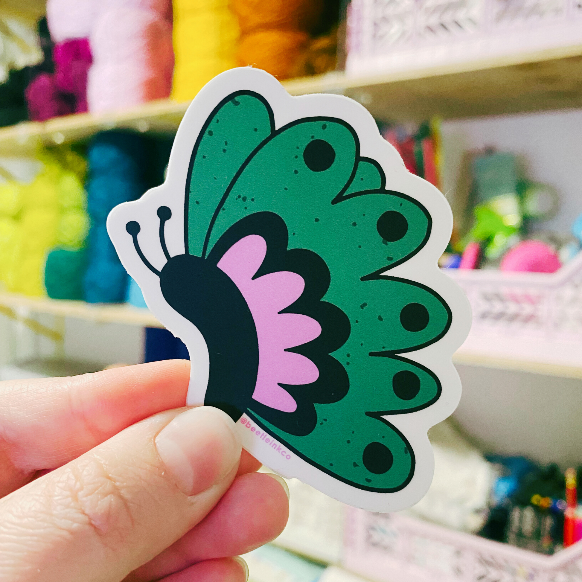 The artist holds up a hand drawn vinyl sticker in the shape of a fluttering butterfly, in her art studio.