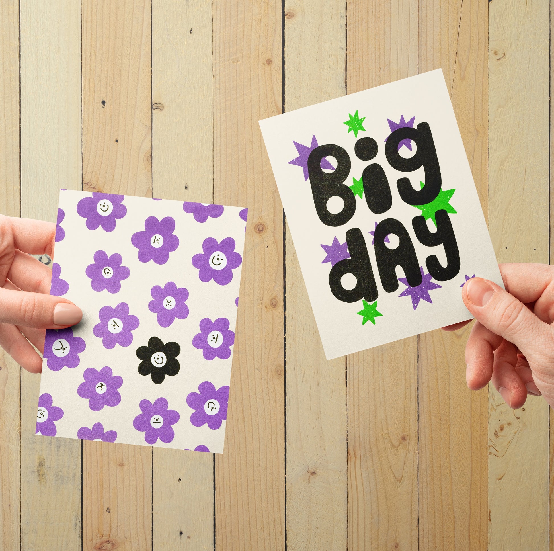 Two hands holding up risograph printed greeting cards, in front of a light wood panel background.
