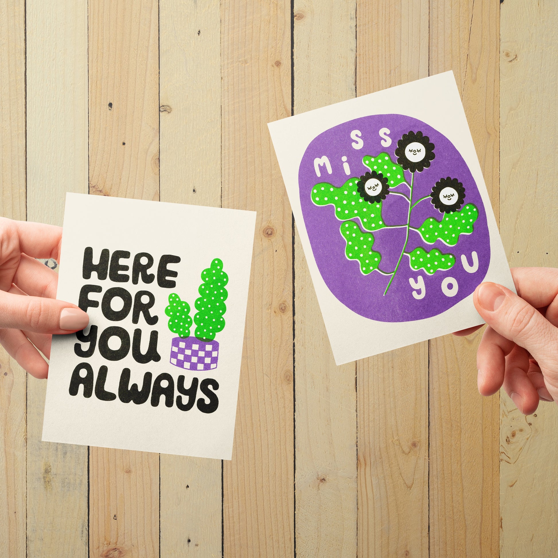 Two hands holding up risograph printed greeting cards, in front of a light wood panel background.