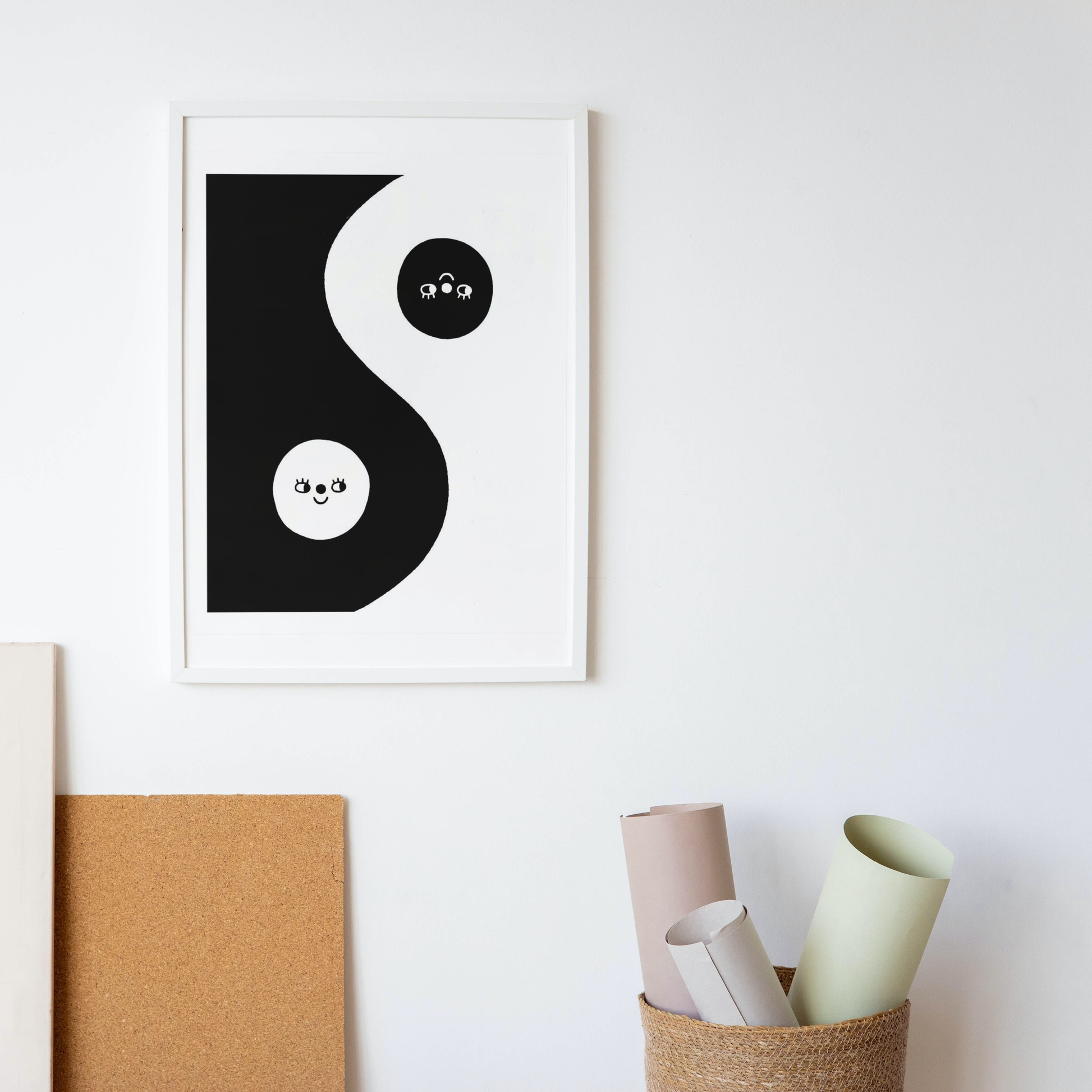 A framed illustrated risograph print of a yin yang symbol with faces, hangs on a white wall with art supplies on the floor below.